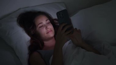 Bir gece yarısı sahnesi: bir kadın, yatağa bağlı, telefonlarının ekranından büyülenmiş, uykusuzluk ve akıllı telefon bağımlılığının zorluklarını somutlaştırıyor.. 