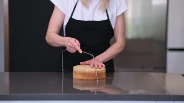 Odaklanmış siyah önlüklü sarışın pastacı modern mutfakta duruyor ve düğün için özel bir şeyler hazırlayan tel bıçaklı yetenekli kadınla hamur hamuru kesiyor.