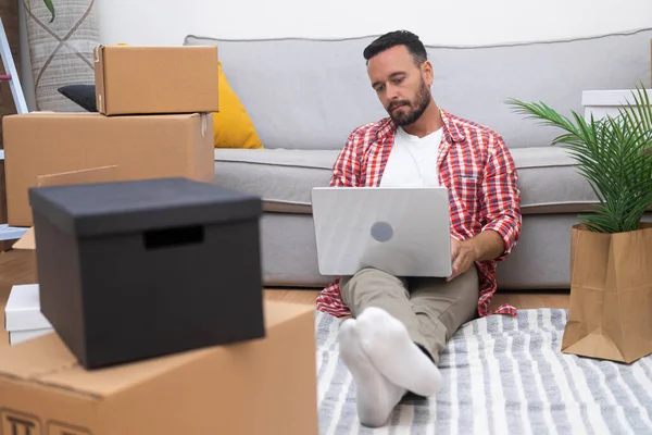 Young man in new apartment with laptop amidst boxes, plans home decor online, setting the stage for a stylish living space
