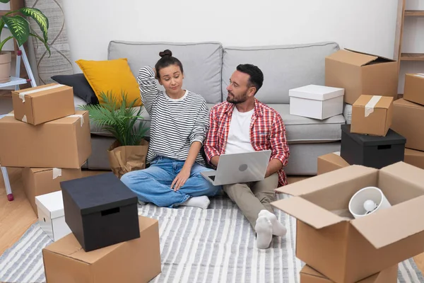 Couple of young people enjoys moving in new modern apartment in another town together sitting with serious expression on floor near sofa among cardboard boxes