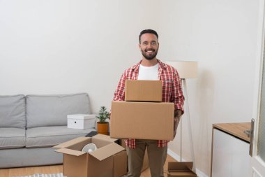 Mutlu adam yeni yenilenmiş oturma odasındaki paketlenmiş nakliye kutularını servis yoluyla taşınmak ve yeni bir daireye taşınmak için eşyalarla dolu olarak tutuyor.