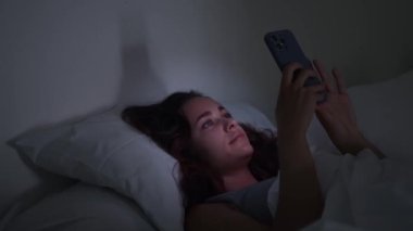 Bir kadın, gecenin karanlığında, yatağa uzanmış, telefonlarına odaklanmış, parıldıyor, uykusuzluk ve akıllı telefon bağımlılığının mücadelesini resmeden bir görüntü.. 