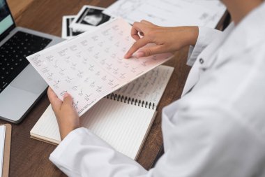 Genç doktor hasta kardiyogram testlerinin sonuçlarıyla gazeteleri okuyor. Kadın kardiyolog, bilgisayar ve steteskopla masada oturan belgeleri inceliyor.