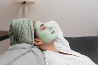 Yüzünde kil maskesiyle rahat bir kanepeye uzanmış, spa prosedürleri sırasında doğal kozmetik ile dinlenen bornozlu güzel bir bayan.