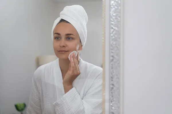 woman, by the bathroom mirror, practices self-care, gently cleansing her skin with a cotton pad.