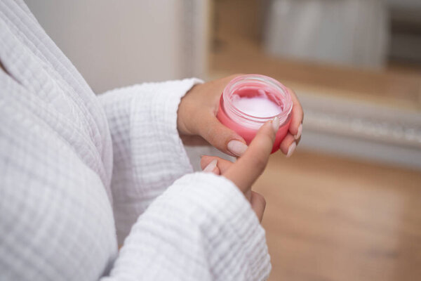 Woman in bathrobe holding open bottle of moisturizing cream near mirror in bathroom lady showing organic cosmetic product for skin pampering in bathroom closeup