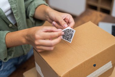 Hassas bir şekilde, çevrimiçi alışveriş yapan bir kadın karton bir pakete QR kod etiketi takarak geri dönüp parasını geri alma niyetini işaret ediyor.. 