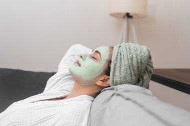 Konforlu Spa: Bir bayan, bornozuyla, yüz nemlendirici maskesiyle rahat bir koltukta gevşer, ev spa tedavilerinin huzurunun tadını çıkarır.. 