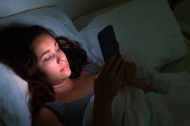 Karanlıkta yatalak bir kadın telefonuna bakıyor. Uykusuzluk ve sosyal medya bağımlılığının yarattığı zorlukların bir yansıması.