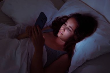 Gecenin sessizliğinde, bir kadın yatağa uzanıyor, gözleri telefonunda, uykusuzluk ve akıllı telefon bağımlılığının sorunlarını vurguluyor.. 