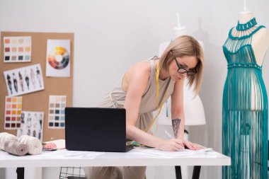 Kadın tasarımcı, terzilik atölyesinde dizüstü bilgisayarla masasında notlar bırakıyor. Terzi, makrome elbisesiyle makrome elbiseler tasarlıyor.