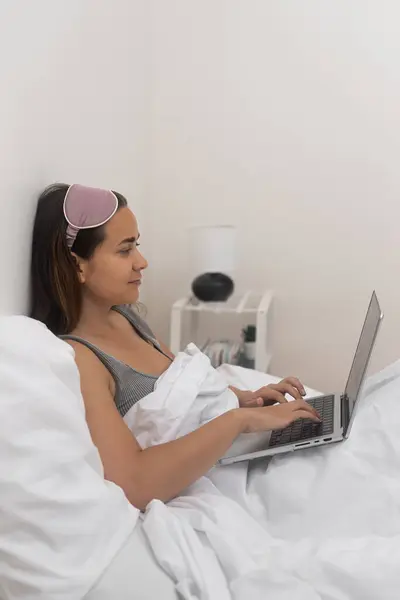 woman in sleep mask works from home in bed, using her laptop to surf the internet and study remotely.