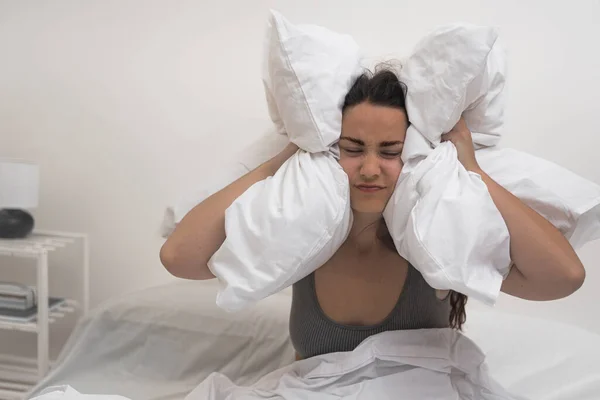 Lady with sleep disorders presses pillows to head frowning young woman experiences migraine attack person suffers from insomnia sitting in bed in light bedroom