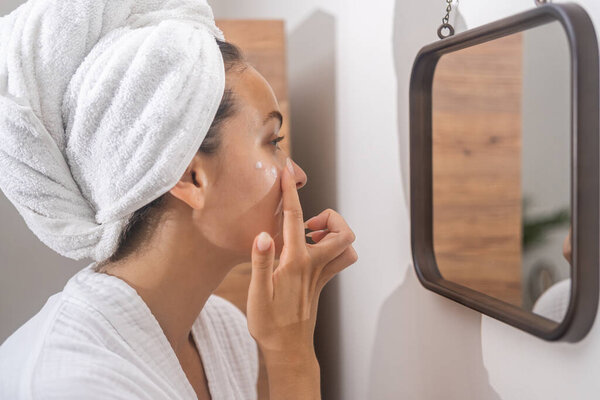 A woman in bathrobe, dedicated to her skincare routine, applies moisturizer in front of the mirror, enhancing her home self-care. 