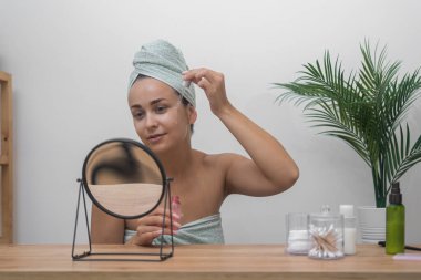 Sakin bir sahne: Kafası havluya sarılı bir kadın, aynalı masasında hiyalüronik asit serumu kullanıyor, rahatlatıcı ev derisini kucaklıyor.. 