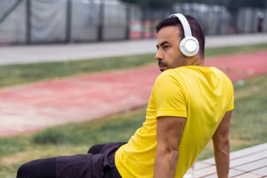 Kablosuz kulaklık takan, ahşap bankta dinlenen sporcu spor sahasındaki antrenmandan sonra mola veren bir sporcu..