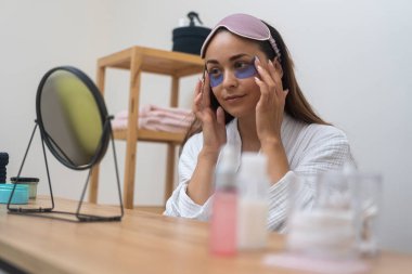 Ev spa düşkünlüğü: Bornozlu bir kadın spa gününde aynaya bakarken yaşlanma karşıtı cilt bakımına odaklanarak göz bandı yapıştırıyor.. 