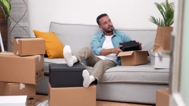 Yeni bir yere taşınma ve yeni bir başlangıç keyfi içinde, genç bir adam taşınma gününde karton kutularla çevrili, yeni evinde mutlu bir şekilde eşyalarını açıyor.