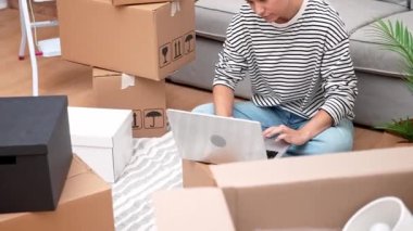 Yeni başlangıçlar: Kadın ev sahibi veya kiracı, kutuların arasında, dizüstü bilgisayarla yerde oturuyor, iç tasarım, çevrimiçi alışveriş veya taşınma hizmetleri hakkında karar veriyor