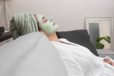 Kanepe Spa: Genç bir bayan, bornozuyla, yüz nemlendirici maskesiyle rahat bir kanepede dinleniyor, spa prosedürlerini uyguluyor.. 