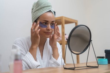 Bornoz güzeli: Bir bayan, yakın planda, bir kaplıca günü boyunca, yaşlanma karşıtı cildini mükemmelleştirirken, göz bantları yapıştırır, yansımasına bakar.. 