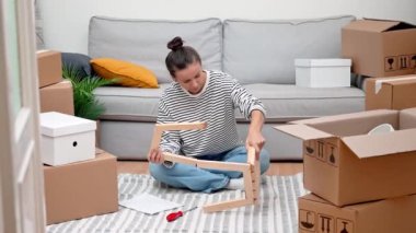 Kira Konforu: Kadın kiracı yerde oturuyor, kendi kendine montaj raflarını karton kutuların ve mobilyaların arasında birleştiriyor, yeni evini evindeymiş gibi hissettiriyor.