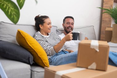 Yeni evde, koltukta oturan genç bir çift paketlenmemiş kutuların arasında kahve fincanlarıyla kutuları açmanın ve yeni bir bölüme doğru heyecanlı yolculuklarının tadını çıkarıyorlar.