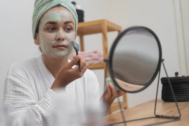 Yüz tazeleme: Bornoza bürünmüş bir kadın cildini organik yüz bakımıyla yükseltir, aynaya bakarken kilden bir maske takarak.