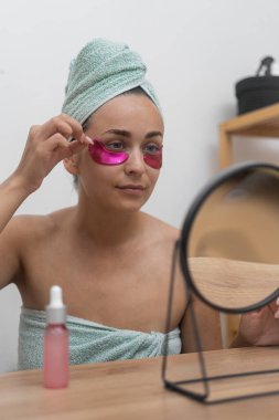 Güzellik sığınağında bir bayan, aynanın karşısında, profesyonel kozmetik ürünlerine düşkündür, cildini geliştirmek için pembe göz altı bantları uygular.. 
