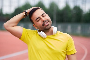 Sarı tişörtlü, boyun tarafı esneyen, ısınma egzersizinde stadyumda atletizm antrenmanına hazırlanan bir adam.