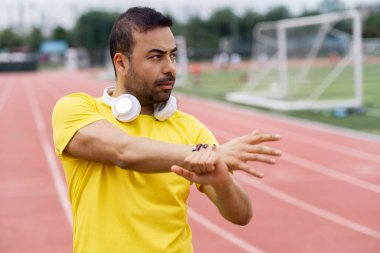 Kulaklıklı düşünceli bir sporcu kol kaslarını ısıtıyor. Spor sahasında antrenman için vücudu hazırlıyor. Yazın futbol sahasında.