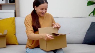 İnternetten sipariş edilen karton kutuları açan gülümseyen kadın, modern apartmanda online alışveriş tecrübesiyle tatmin olmuş bir şekilde kanepede oturuyor.
