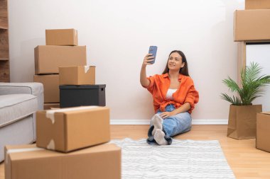 Genç bir kadın elinde telefonuyla satın alınmış evdeki büyük paketlerin arasında oturuyor. Sevdikleri için çağdaş evlerde taşınırken hatırlanmaya değer fotoğraflar çekiyor.