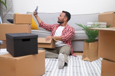 Yakışıklı adam cep telefonunu elinde tutuyor. Yeni bir dairede çağdaş bir fotoğraf çekiyor. Koltuğun yanında, halı döşenmiş, büyük paket kutularının arasında oturuyor.