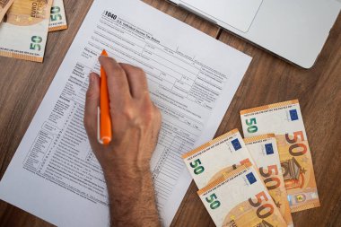 Tüccar, vergi hesaplamak için dijital varlık işlemlerinden elde edilen kazanç ve kayıpları rapor eden masada avro banknotları ve dizüstü bilgisayarla çevrili 1040 formu dikkatlice dolduruyor