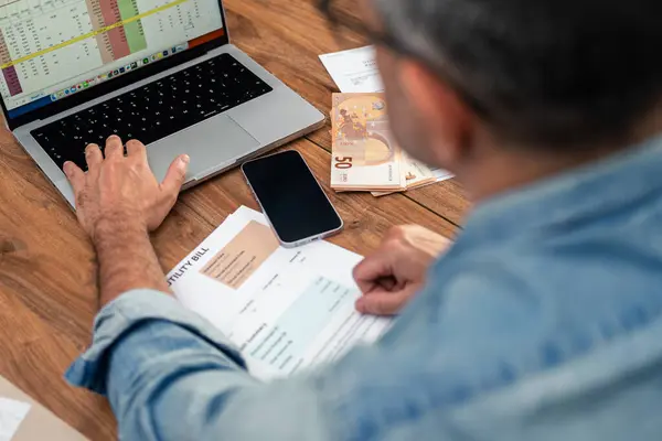 Mavi gömlekli adam faturaları inceliyor ve masadaki para ve akıllı telefonla dizüstü bilgisayardaki masrafları hesaplıyor. Ev yönetimi ve çevrimiçi ödemeler için mali planlama ve bütçe 