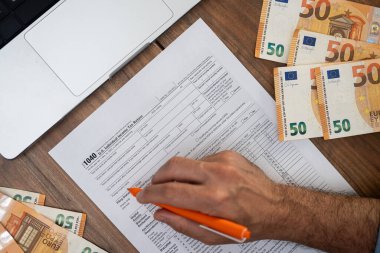 Hand of a man filling out a 1040 tax form with euros and a laptop on a wooden desk. Tax preparation and financial documentation concept, suitable for accounting and international finance themes  clipart