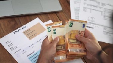 Orta yaşlı bir adam, ev idaresi faturalarıyla ilgili vergi formlarının yakınındaki 50 avroluk banknotları dikkatle sayıyor.