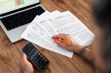 Erkek şirket muhasebecisi, iş yerinde dizüstü bilgisayarla oturan doğru mali veriler için akıllı telefondaki hesap makinesini kullanarak vergi formlarını doğru şekilde dolduruyor.