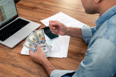 Masada ev giderleri üzerinde çalışırken elinde dolar tutan bir adamın yakın çekimi. Finansal planlama, bütçe, faturalar ve bir dizüstü bilgisayar. Ev finansmanı ve ödeme yönetimi kavramı 