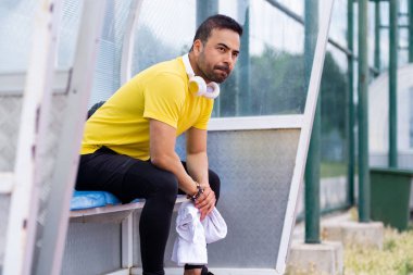 Resting in the arbor of the city sports stadium, the bearded sportsman, wearing headphones and with a towel nearby, enjoys a moment of tranquility on the outdoor athletic arena.  clipart