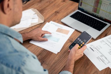 Olgun işadamı fatura numaralarını ofis masasında akıllı telefon uygulaması ile ekliyor, üst düzey erkek çalışan ev giderlerini ödemeye hazırlanıyor, faturalar belgesi