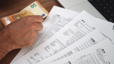 İşadamı, avro banknotlarını elden ahşap masaya aktararak para sayıyor, faturaları vergi beyannameleri ve dizüstü bilgisayarların yanına koyuyor