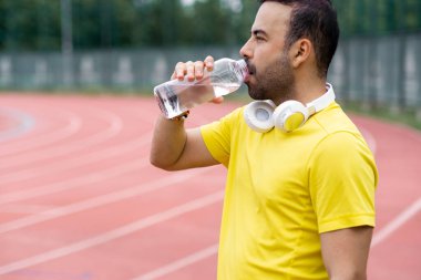 Hidratlaştırma ve şarj etme, kablosuz kulaklıklı sporcu spor sahasındaki koşu pistinde serinletici bir yudum alır ve dengesini korur..