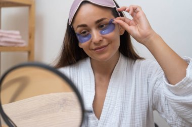 Ayna Bakışı: Ayna yansıması, şişkinlik, kırışıklıklar ve göz altı maskeli koyu halkaları ele alırken bir kadının memnuniyetini yakalar. 