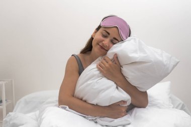 Yatak odalarında sabah parıltısı, uyku maskeli büyüleyici bir kadın sırıtıyor, pofuduk beyaz bir yastık tutuyor. 