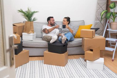 Heyecanla, genç bir çift kahve fincanlarını kanepeye kaldırır, paketsiz kutuların arasında, kutuları açarken ve yeni evlerine yerleşirken..