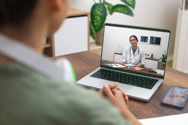 Over-the-shoulder view of a patient having a video call with a smiling doctor on laptop. clipart