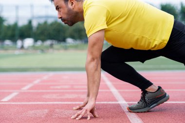 Düşük başlangıç pozisyonunda odaklanmış sporcu, bir şehir spor sahasının canlı kırmızı kauçuk pistinde koşmaya hazırlanıyor. Açık havada yoğun bir idman..