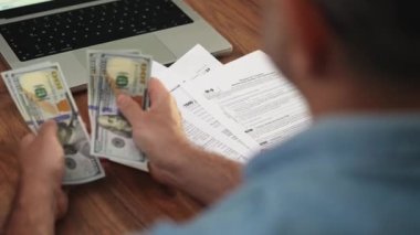 Vergi formlarını doldurmadan önce adam dolarları elden ele aktararak para sayıyor, ofis çalışanı masası dizüstü bilgisayarla düzenleniyor ve beyannameler yavaş çekimde düzenleniyor.